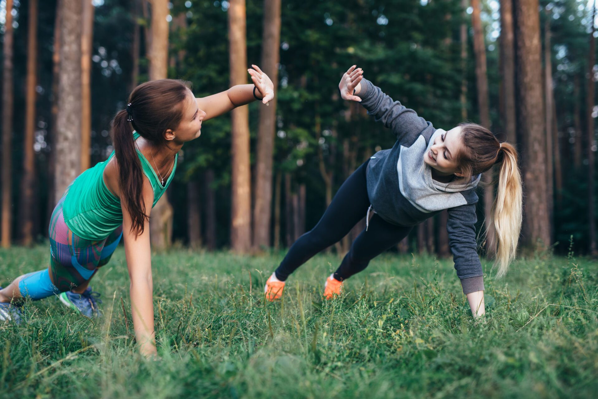Two female buddies doing partner side plank giving high five while training in the forest.
